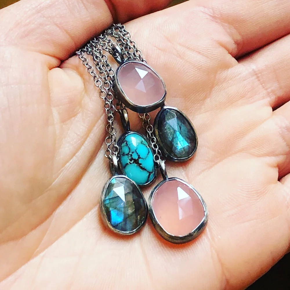 Hand holding a silver chain necklace with five oval-shaped gemstone pendants in pink, turquoise, and blue-green hues.