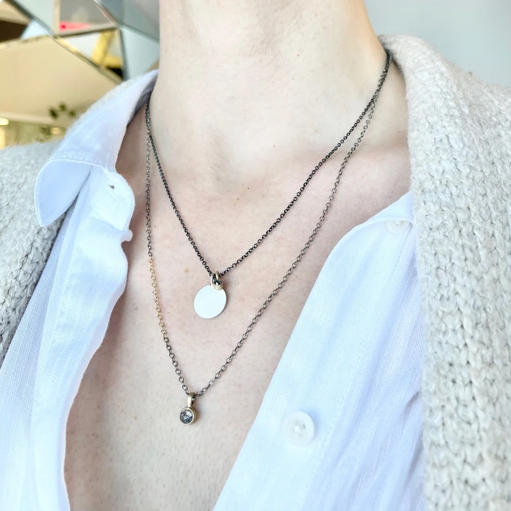 Close-up of a woman wearing layered necklaces with circular pendants, a white collared shirt, and a beige textured coat.