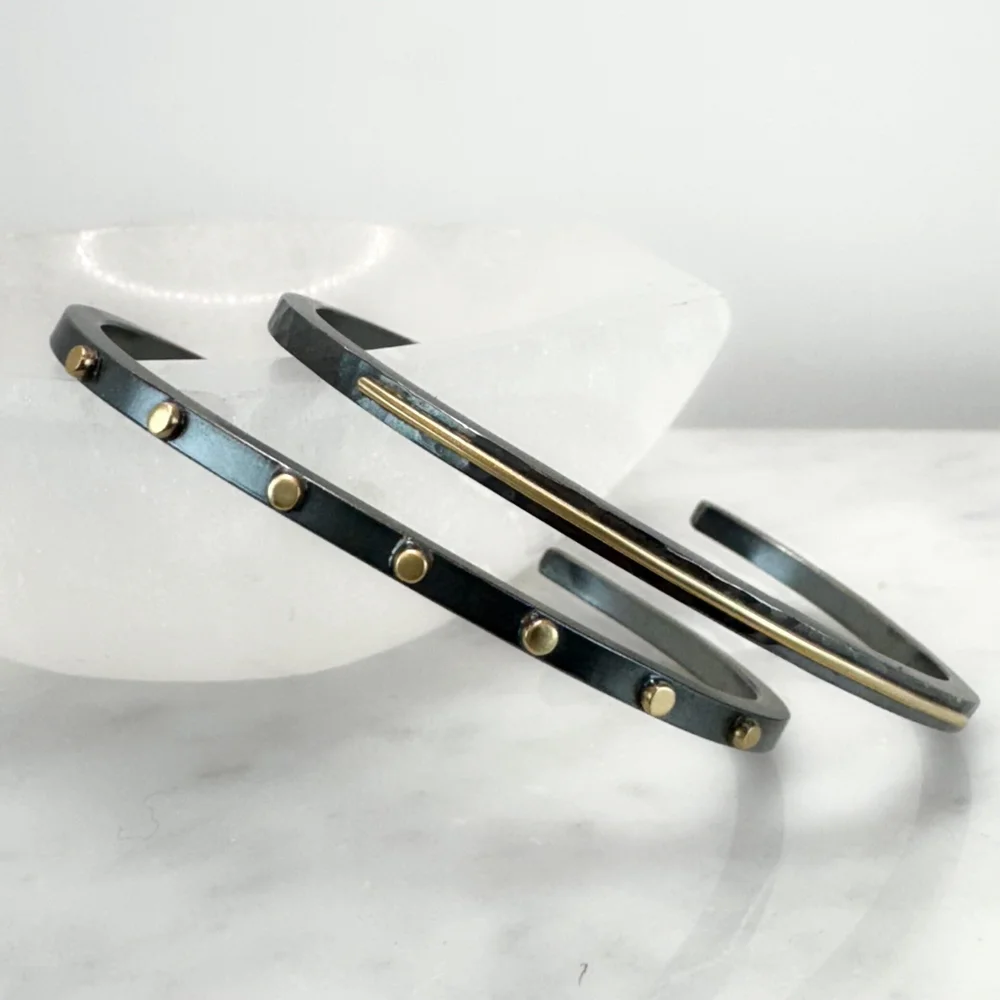 Close-up of two oxidized sterling silver and 14 K gold metal cuff bracelets with small gold balls and a thin gold stripe, resting on a reflective white surface.