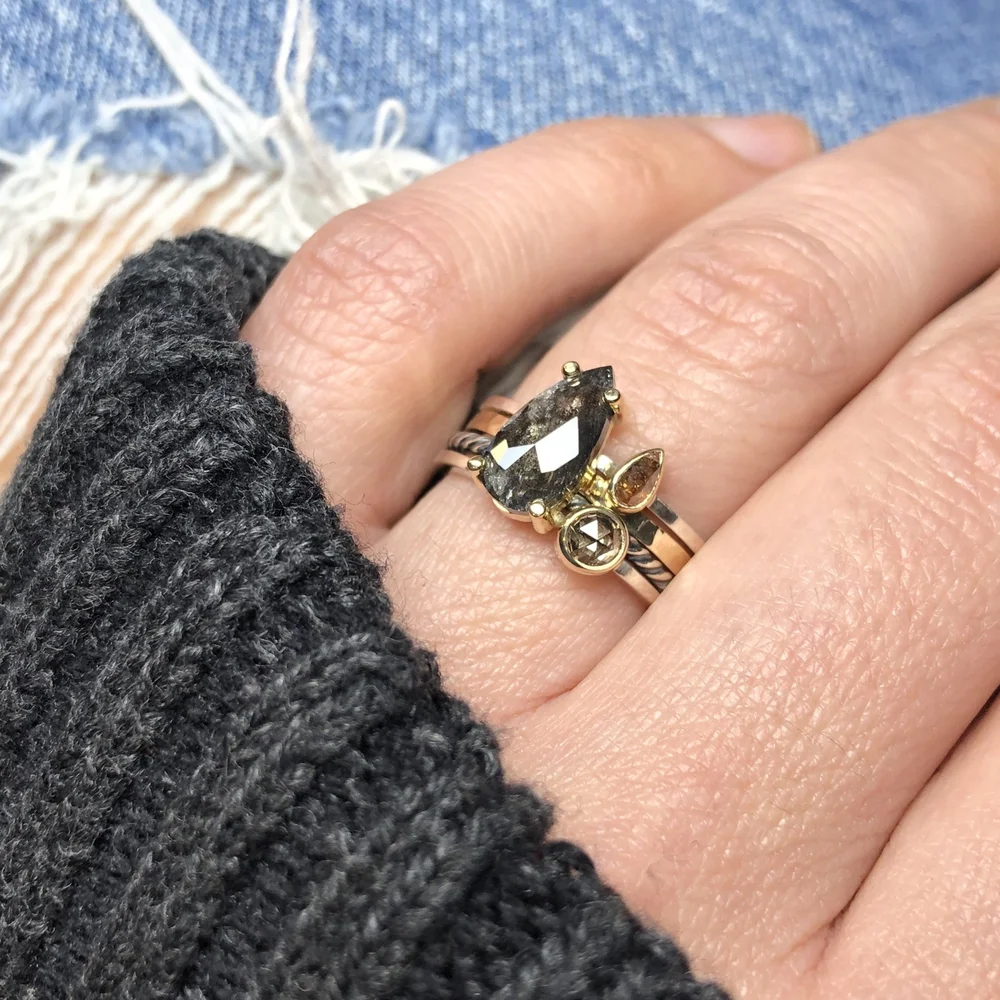 Close-up of a hand wearing two rings, one with a large black gemstone and smaller accent stones, and a black knitted glove, with a denim background.