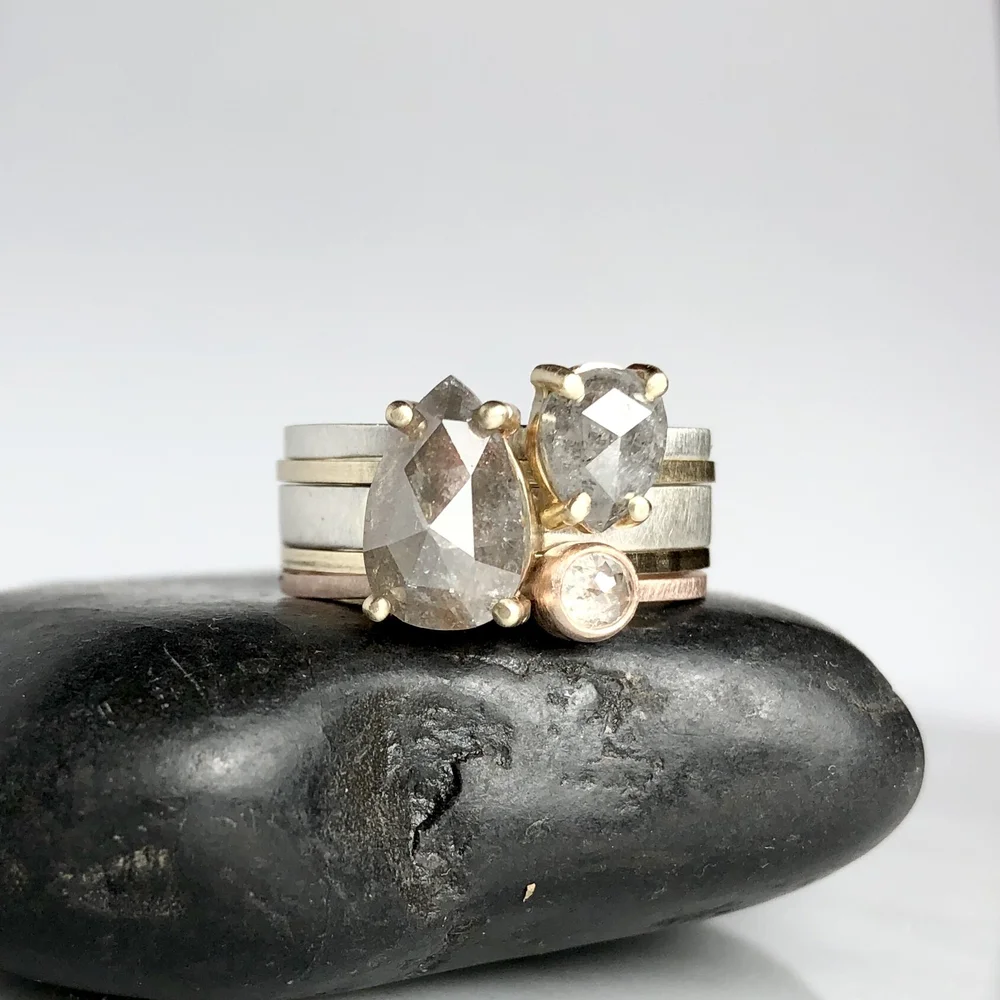Stack of sterling silver and 14K gold rings adorned with three salt and pepper diamonds, two large pear-shaped and one small round, resting on a black stone against a plain white background.