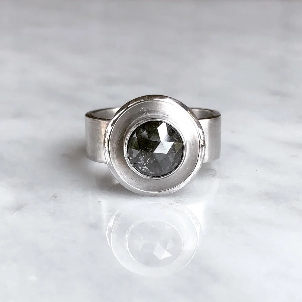 A silver ring with a large, round black gemstone in the center, reflecting light, on a light-colored surface.