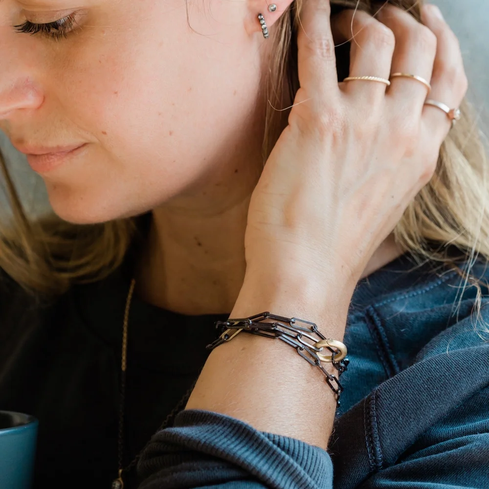 A woman with light skin and blonde hair, wearing multiple rings on her fingers, a layered chain necklace, and a jewelry bracelet on her wrist. She is touching her ear and wearing a black top or jacket.