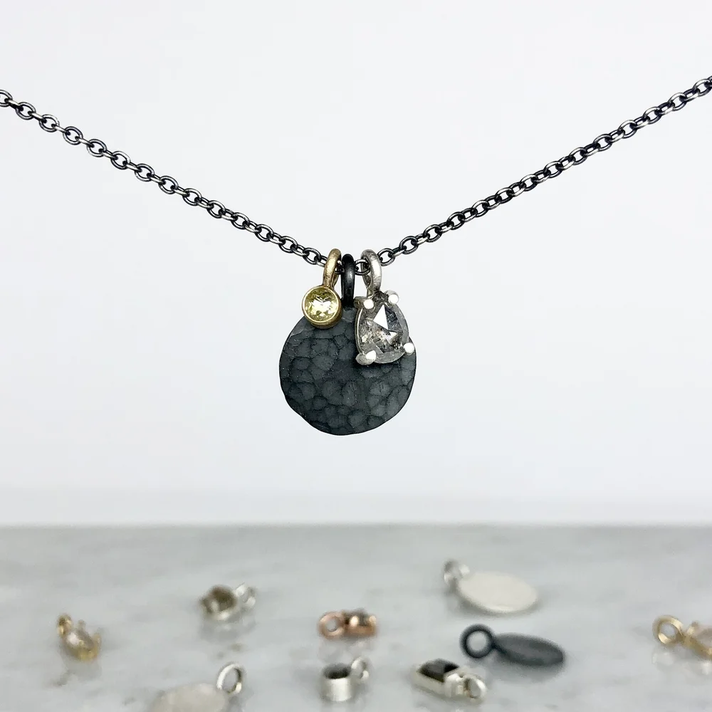 A necklace with two charms, one black textured circular pendant and a clear gemstone, hanging on a chain. Small metal jewelry pieces are scattered below on a light surface.