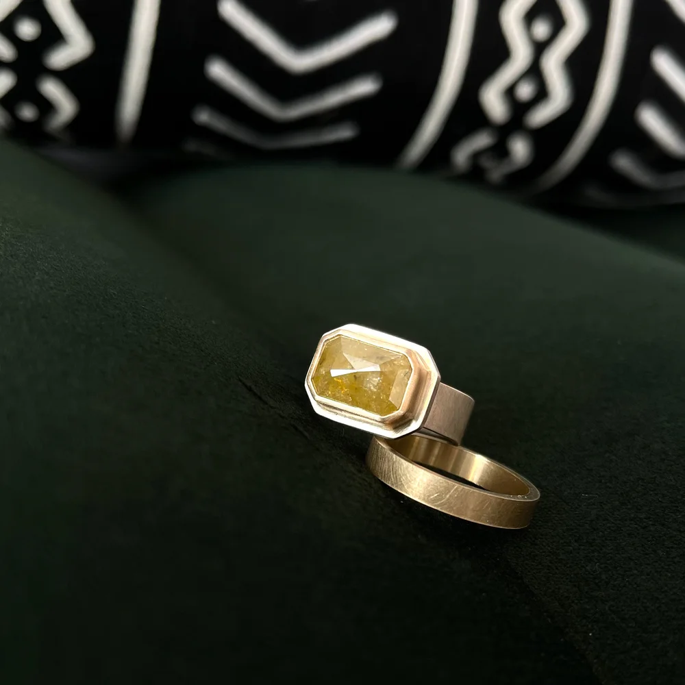 A gold ring with a rectangular yellow gemstone, resting on a dark green surface with a blurred black and white patterned fabric in the background.