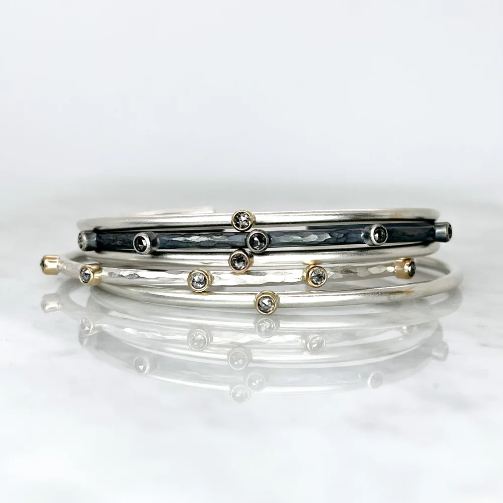 Three sterling silver cuff bracelets with small round salt and pepper diamonds set in bezels, arranged on a reflective white surface.