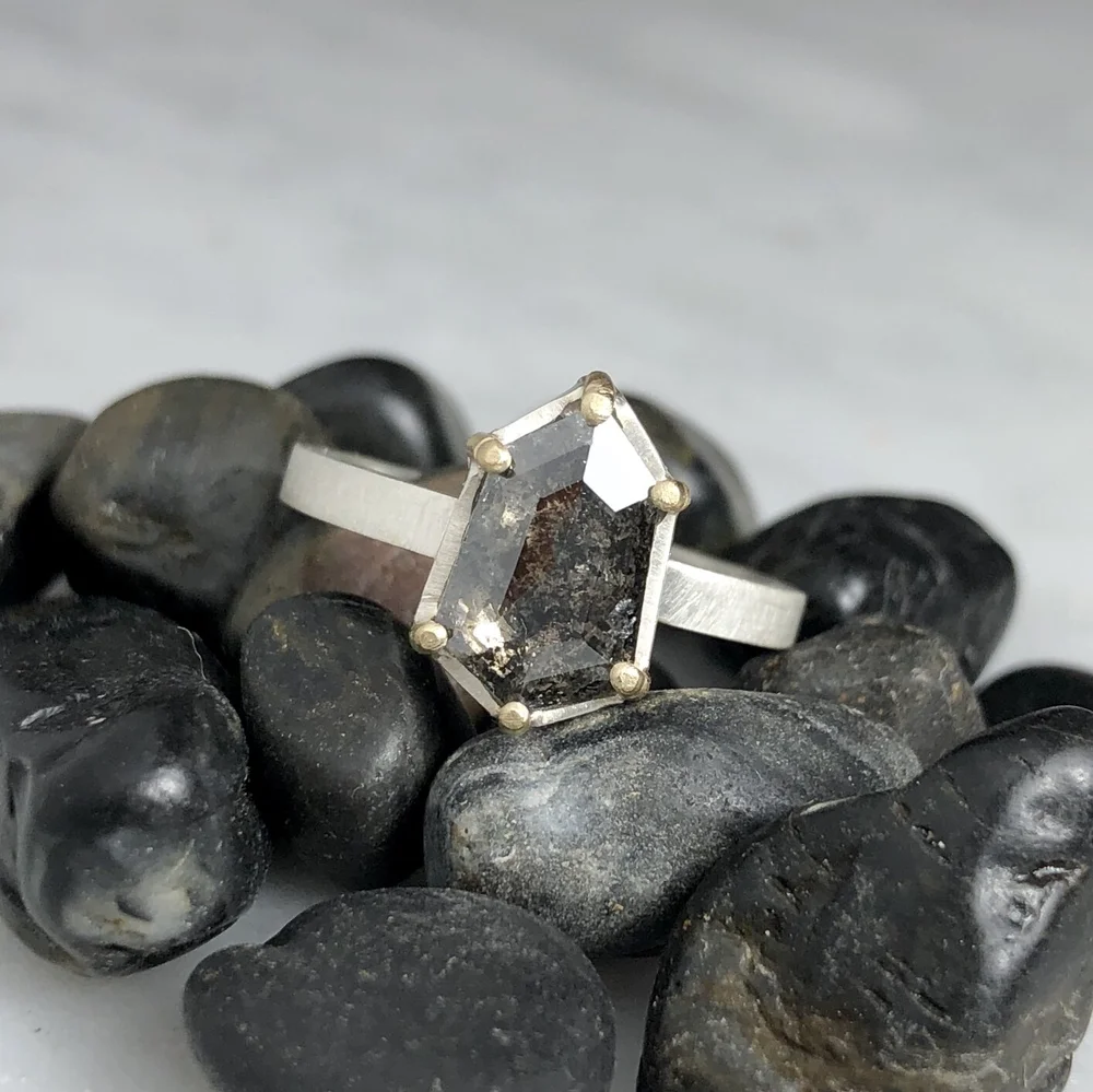 A silver ring with a hexagonal black stone set in a prong setting, resting on black pebbles.