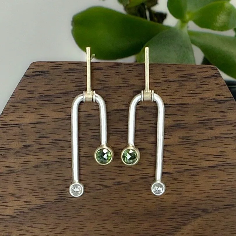 A pair of silver and gold earrings with green and clear gemstones displayed on a wooden stand, with green leaves in the background.