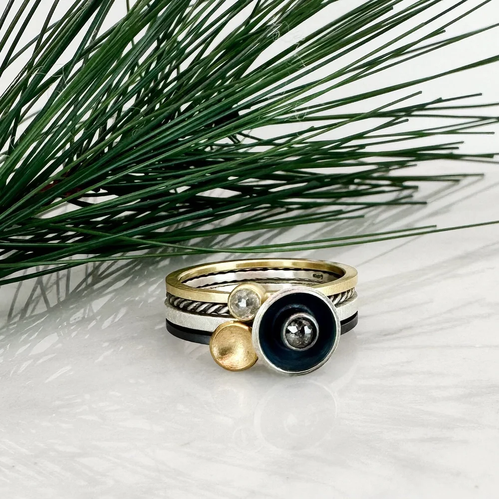 Stacked rings with a black, silver, and gold design, placed next to green pine branches on a white surface.