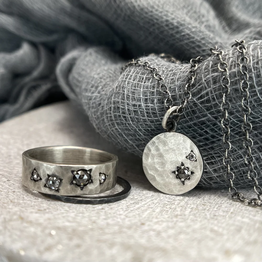 Silver jewelry set including a ring with small embedded stones and a pendant necklace, displayed on a textured fabric background with a grey and black cloth.