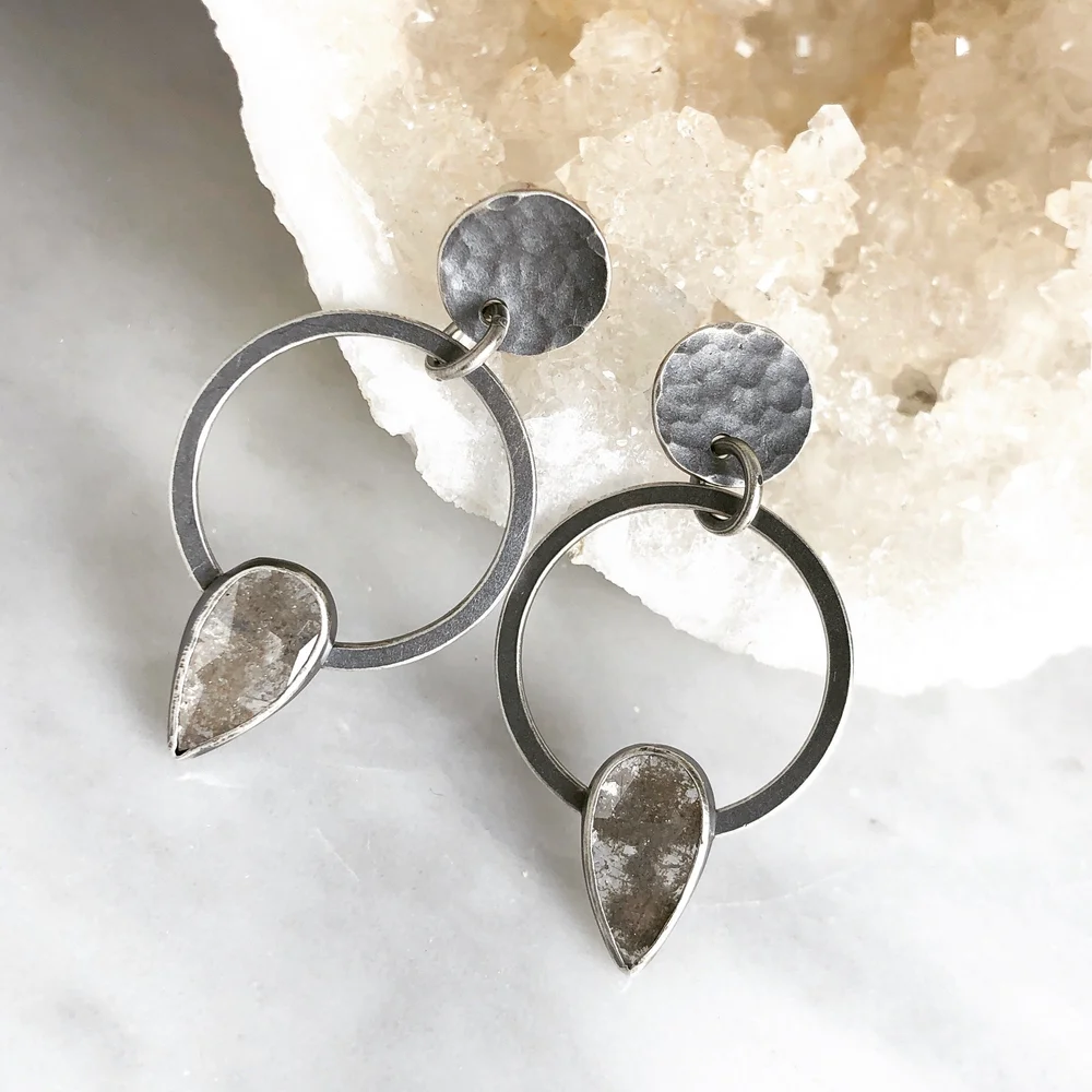 Pair of silver drop earrings with hammered texture and teardrop-shaped pendants, placed on light-colored marble surface with a salt rock in the background.