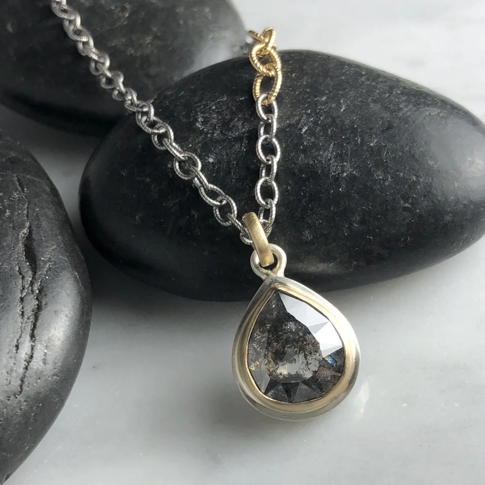A sterling silver and 14K yellow gold necklace with a pear-shaped pendant containing a salt and pepper diamond, resting on black stones.