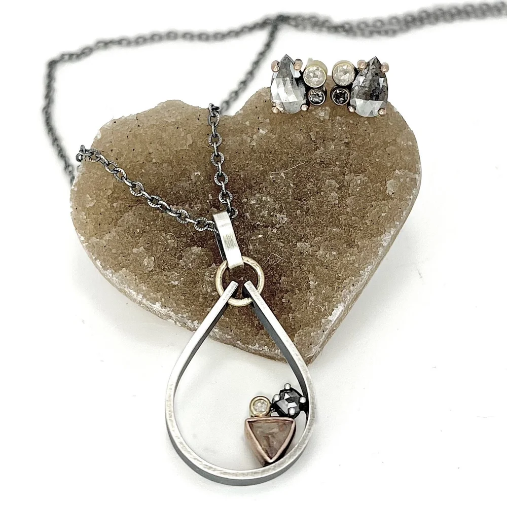 Set of jewelry including a necklace with teardrop pendant and a pair of earrings, placed on a brown heart-shaped stone. The necklace has a circular pendant with various small stones inside, and the earrings feature large, dark and light-colored stone