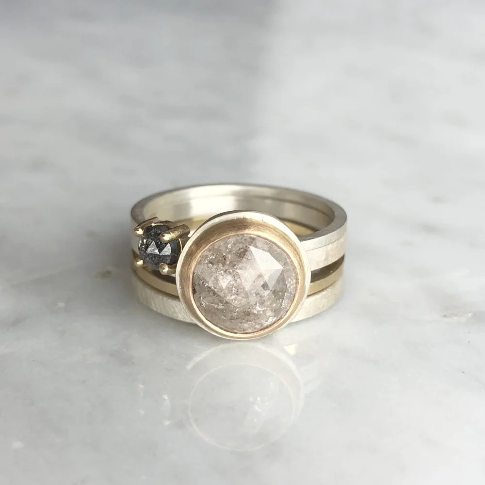 Three stacked rings on a white marble surface: a gold ring with a large round transparent stone, a silver band, and a gold ring with a small dark gemstone.