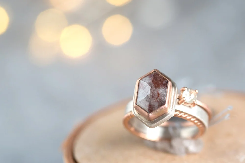 A close-up of a stack of rings on a wooden display stand. One ring features a sterling silver band with a large hexagonal rustic red salt and pepper diamond, and the other has a small round icy white diamond on a 14K rose gold band.