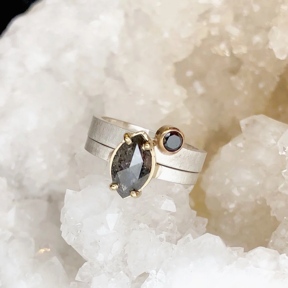 A close-up photo of two rings with black gemstones resting on a bed of white crystalline minerals. The larger ring features an oval-shaped black gemstone set in a gold bezel, and the smaller ring has a round black gemstone set in a gold bezel.