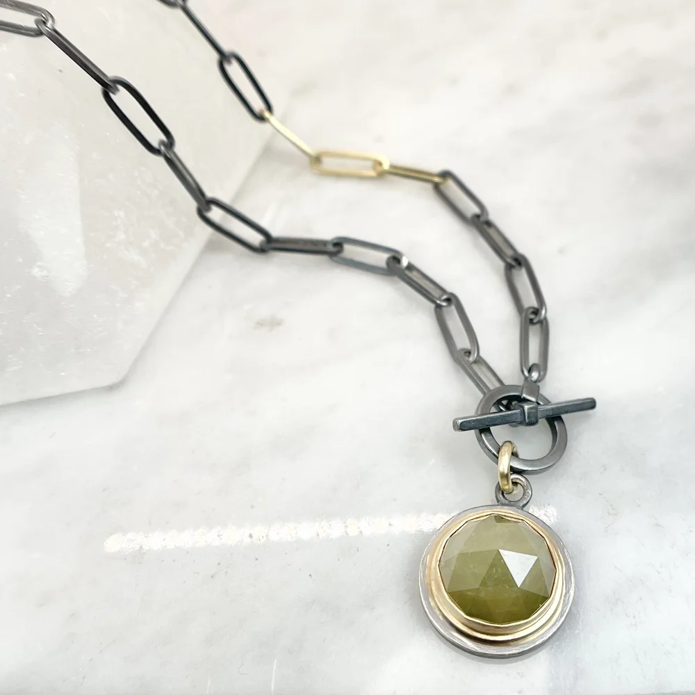 A necklace featuring large black chain links, a gold chain section, and a pendant with a large faceted yellow gemstone. The pendant is attached via a black ring and a gold ring, with a black horizontal bar through a smaller circle.