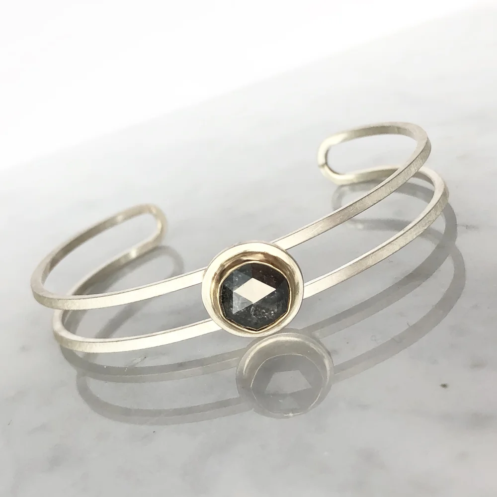 Silver bracelet with a central black gemstone set in a gold bezel, on a reflective surface.